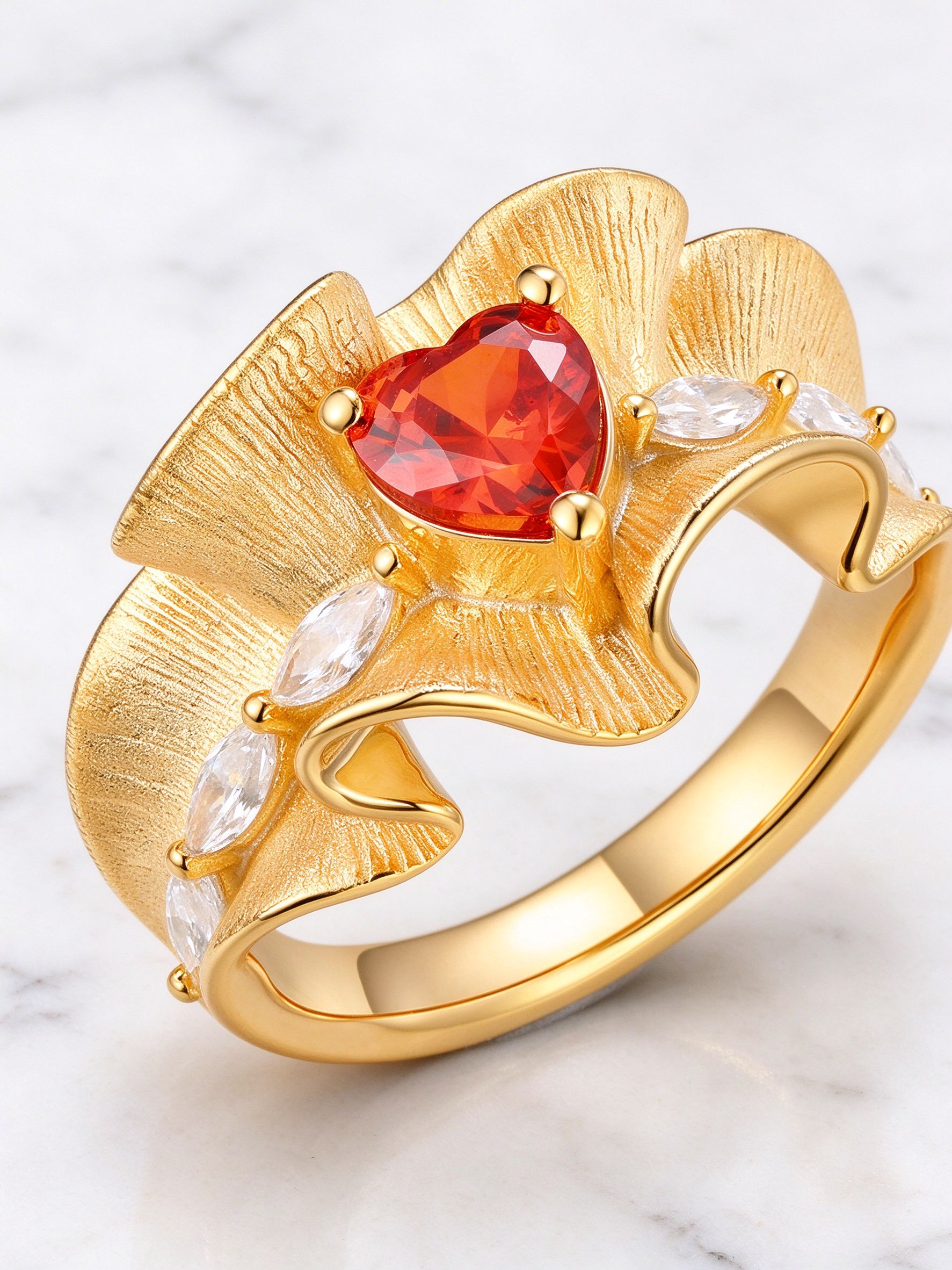 Material detail of brushed gold ruffle band, heart red CZ and marquise clear accents on white marble